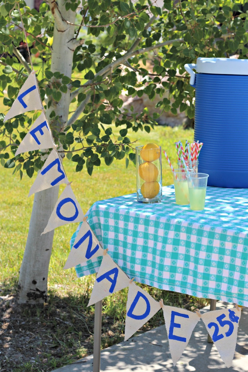 hoto of a Lemonade stand, that has a banner with the word lemonade on it, a green checkered tablecloth on a card table, several glasses of lemonade on the table plus a blue igloo cooler.