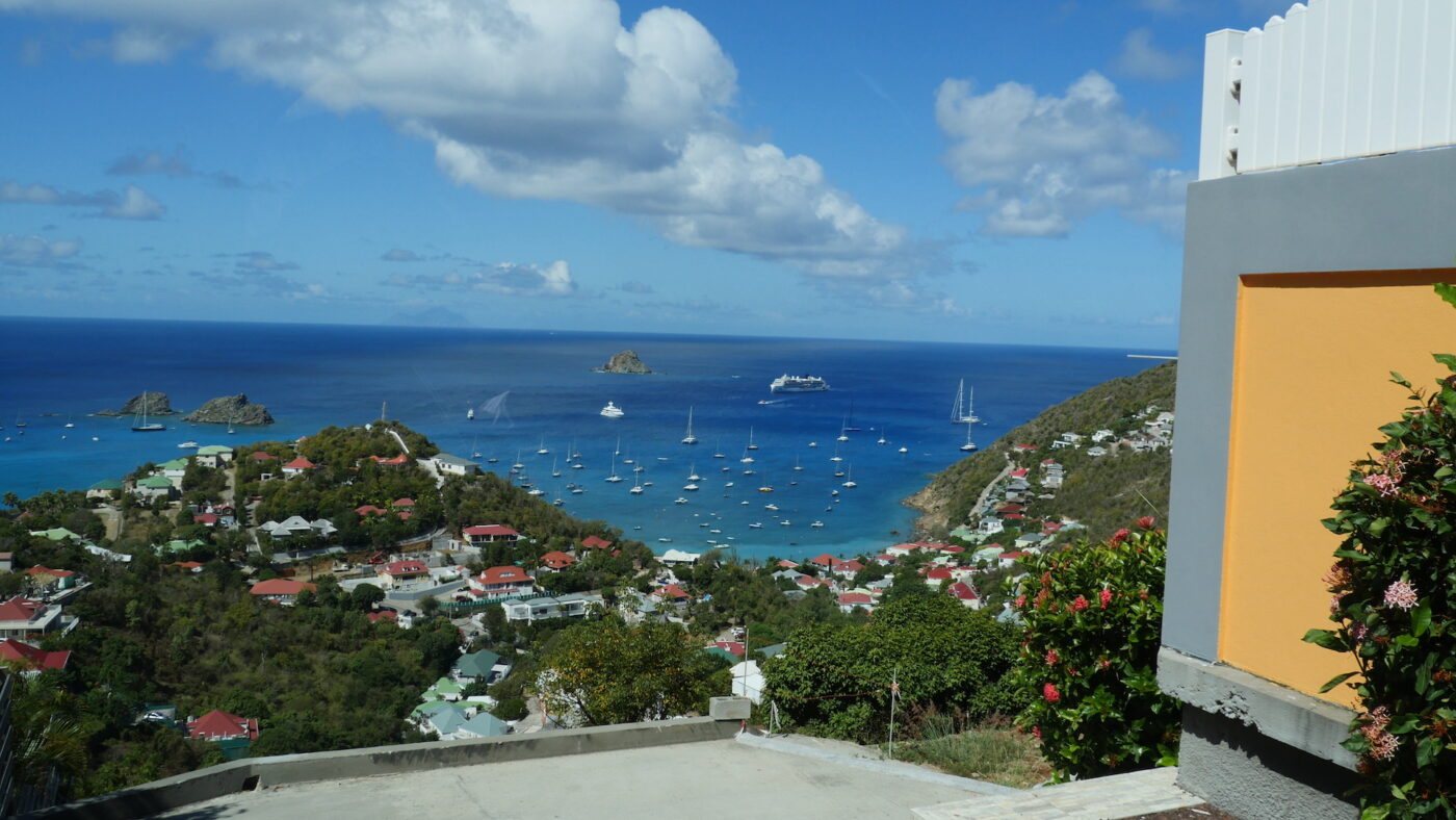 Vista of Caribbean island from top of unknown island. April, 2022.IN foreground are Hillside homes, green foliage with sailboats in the bay, and ocean and clouds beyond.