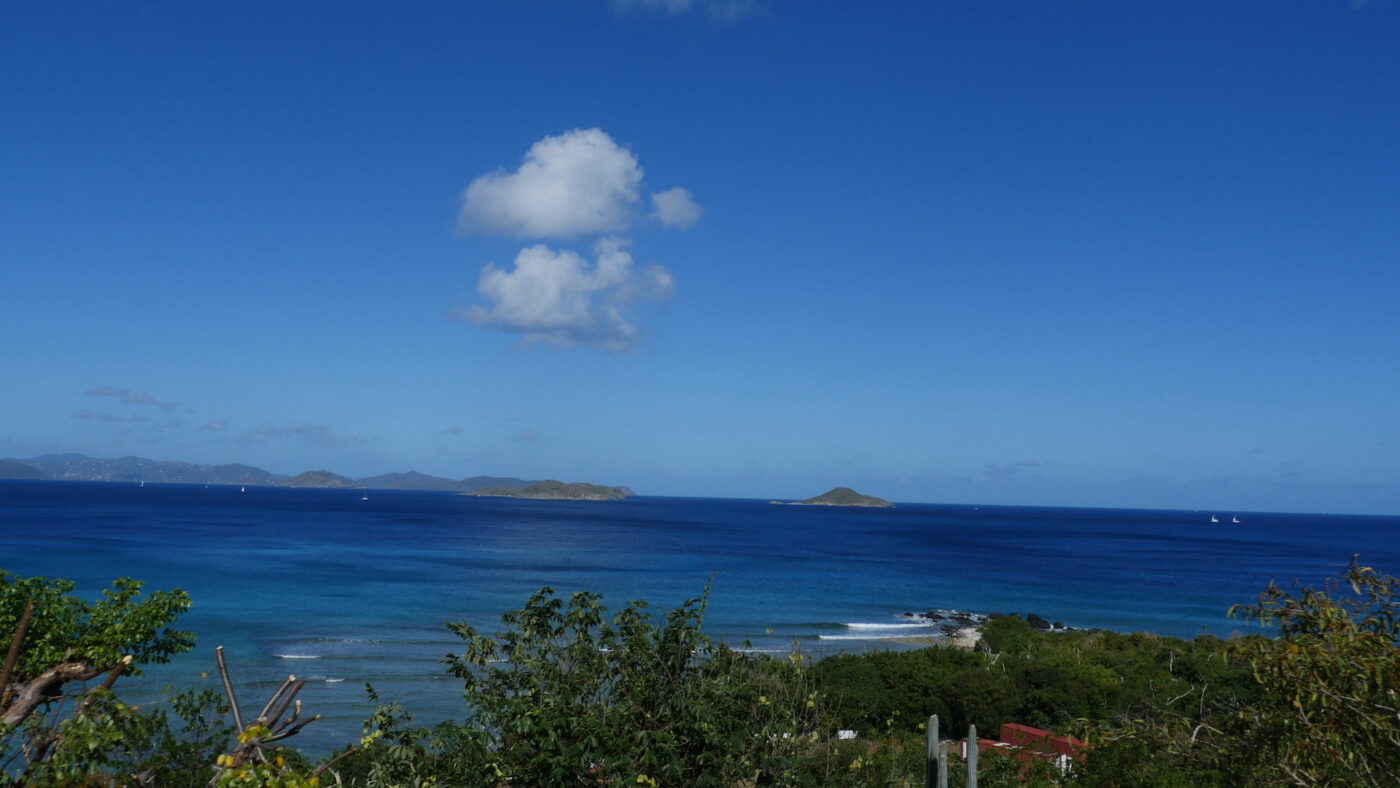 Vista of Caribbean from a hilltop with green foliage in foreground of an unknown island. April, 2022. Beyond is the blue ocean and another island several miles away.