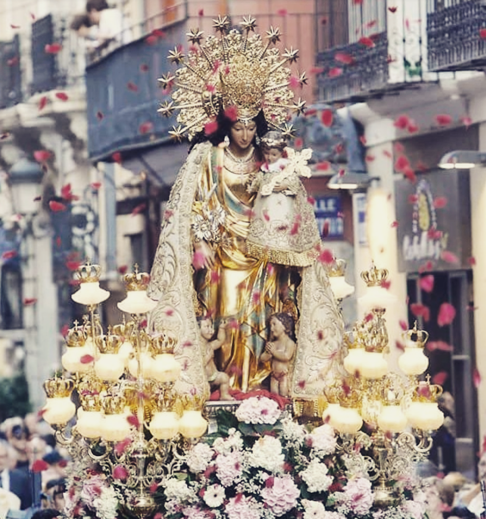 A photograph of the miraculous statue of Our Lady of the Forsaken in a parade procession. Her head is slightly tilted as is baby Jesus. The statue is adorned with beautiful flowers. Photo credit is for All About Mary on Tumbler.