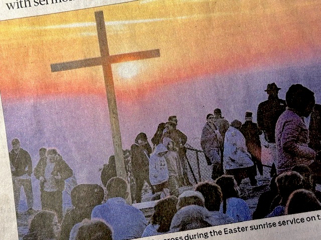 Photo of the March 31, 2024 sunrise from a mountain top. In the foreground is a 20 foot wooden cross and pilgrims with coats and blankets gathered for the Easter Sunday sunrise service at Stone Mountain, Georgia. Photo from the April 1st AJC front page.
