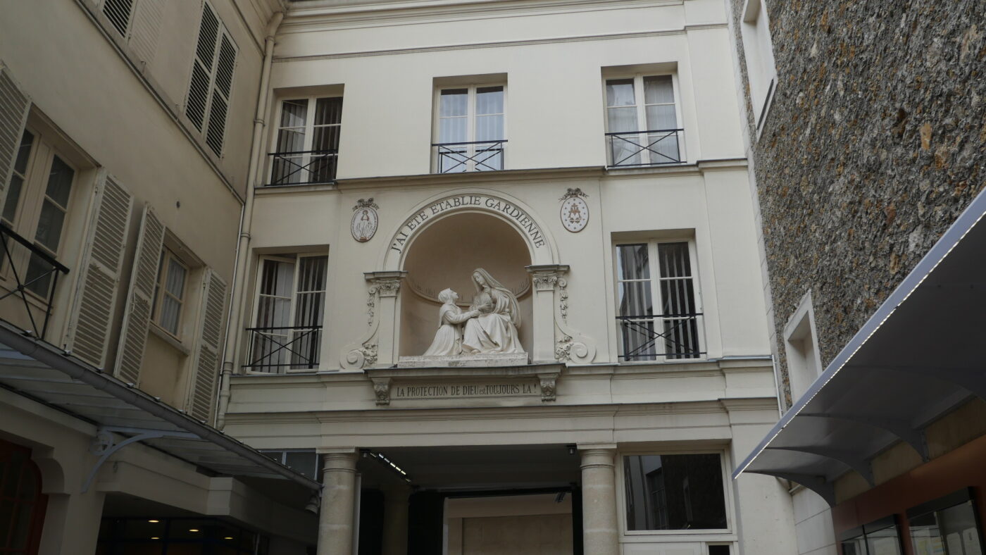 Photo of the building that houses the Chapel of Our Lady of the Miraculous Metal on Rue de Bac, in Paris. The modest ivory facade is on the interior of the humble building. It is a 3 story building with windows on the top 2 floors. In the center of the picture is a sculpture of The Virgin Mary talking to Catherine Labouré who is kneeling next to her.